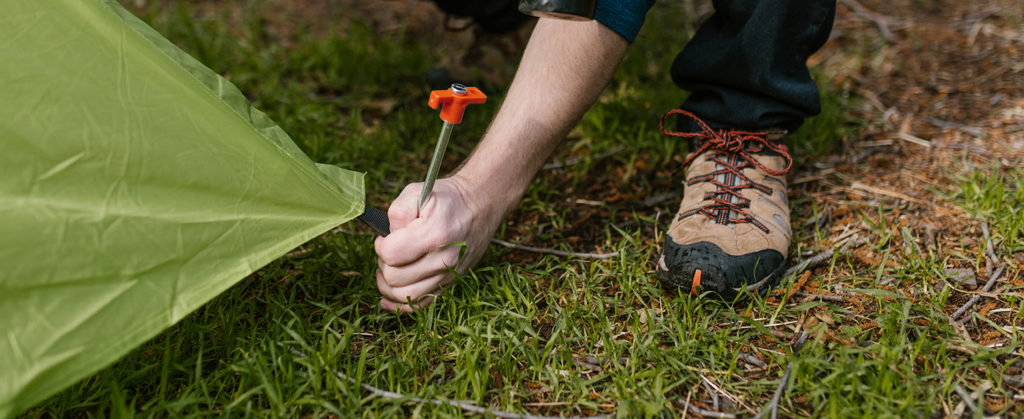 Person securing tent with a stormpløkker on grassy ground.